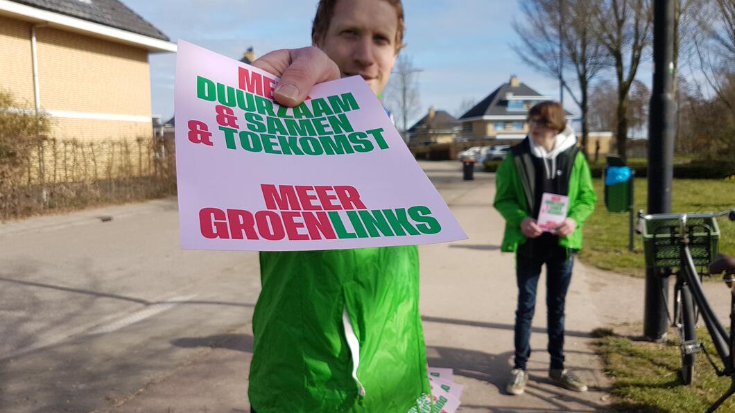 Een vrijwilliger houdt een flyer van GroenLinks in beeld met een woonwijk op de achtergrond.