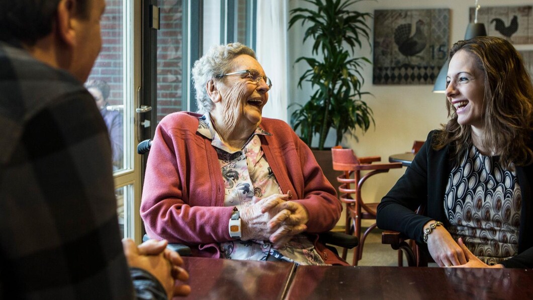 Een oudere vrouw en een jonge vrouw zitten aan tafel en kijken elkaar lachend aan.