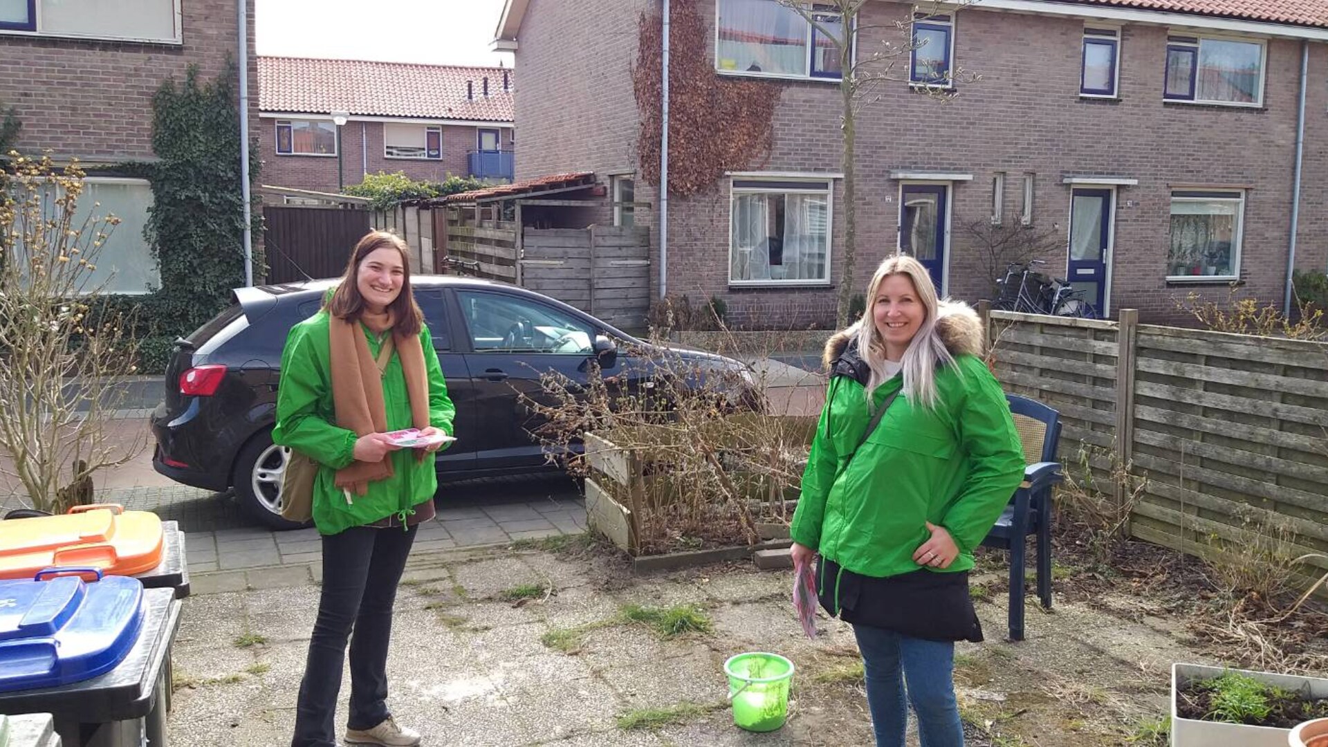 Twee vrijwilligers staan in groene jasjes in een woonwijk.